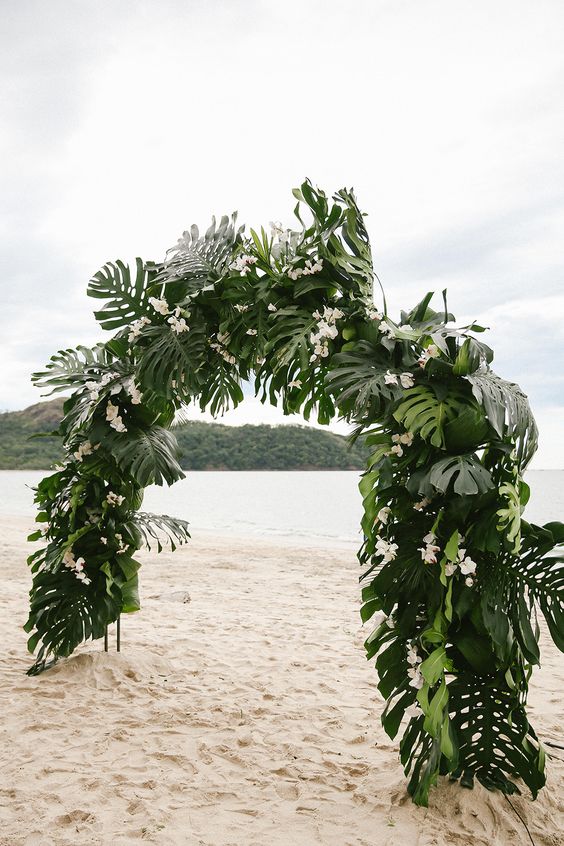 a lush tropical wedding arch covered with fronds and some white orchids is a pretty idea for a tropical beach wedding