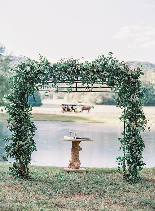a metal wedding arch decorated with lush greenery and with a view of the lake is a lovely idea for an outdoor wedding