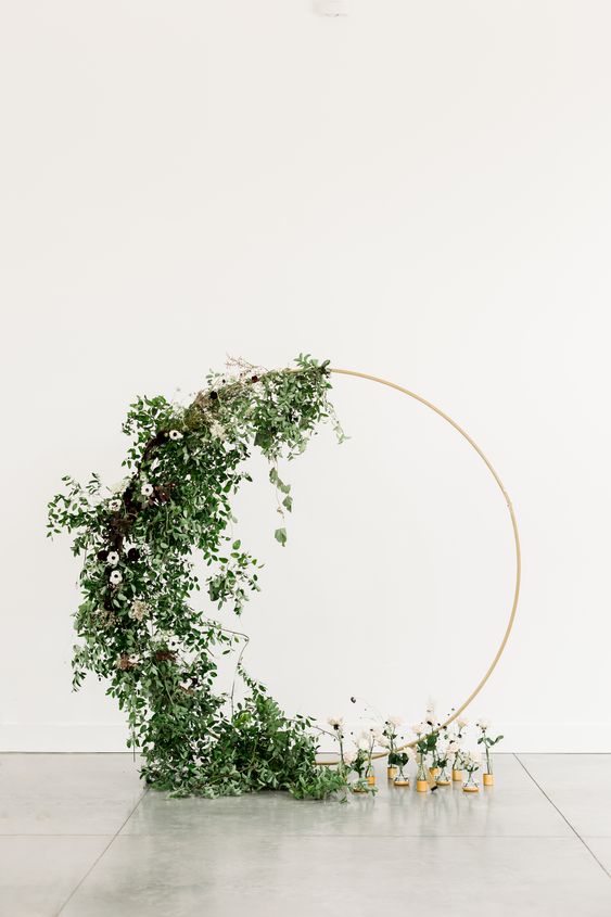 a modern to minimal round wedding arch partly covered with greenery and some white blooms for a modern wedding