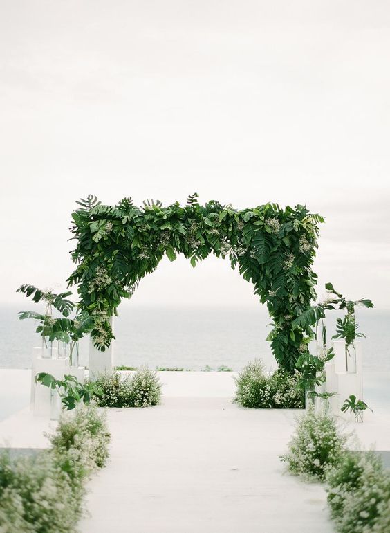 a modern tropical wedding arch covered with leaves and fronds and with greenery along the aisle is amazing for a modern tropical wedding