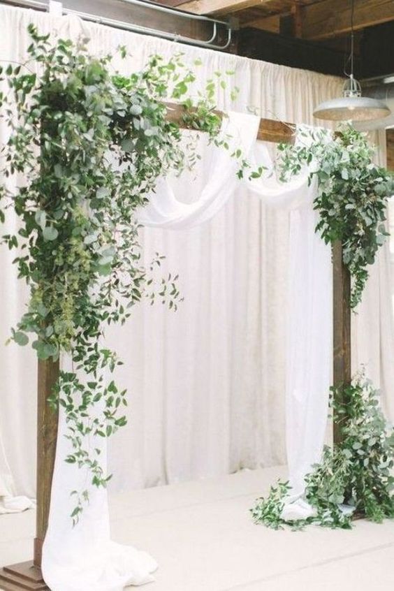 a modern wedding arch covered with greenery and white fabric is a cool idea for a spring or summer wedding