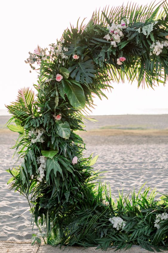 a tropical round wedding arch with leaves and fronds and some blush blooms is a cool idea for a tropical wedding