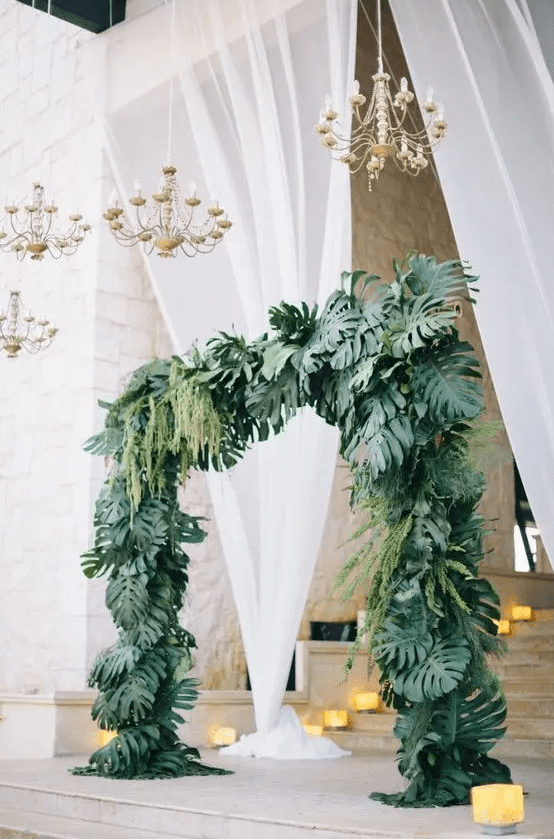 a tropical wedding arch covered with tropical leaves and hanging greenery plus glam chandeliers over it