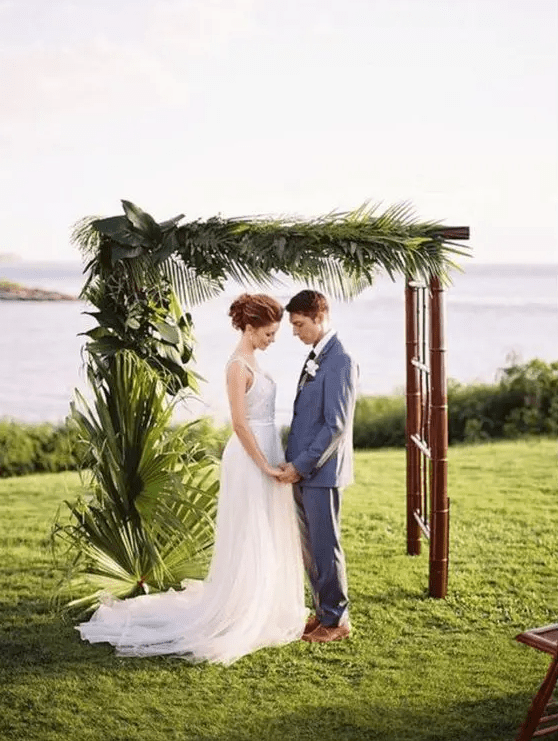 a tropical wedding arch with lush tropical greenery on one side is a cool idea for a tropical wedding, and a sea view adds to the space