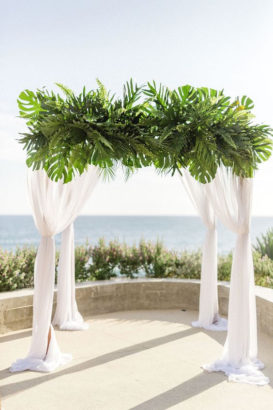 a wedding chuppah with white curtains and fronds on top is a cool idea for a modern tropical wedding