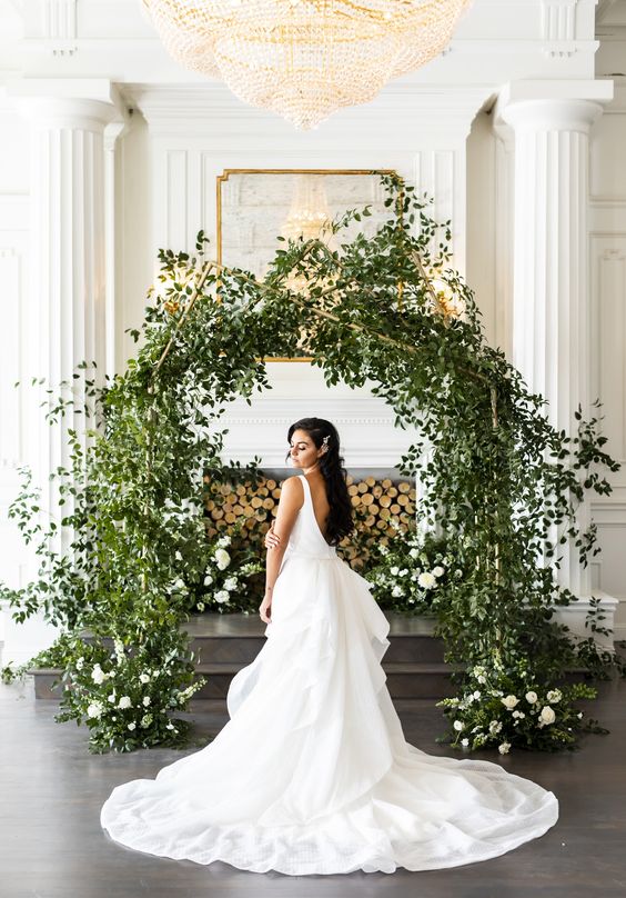 an indoor greenery wedding arch of a peculiar shape with some white blooms creates a feeling of outdoors