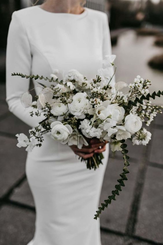 a modern winter bridal look with a plain fitting wedding dress with a high neckline and long sleeves plus a white ranunculus wedding bouquet