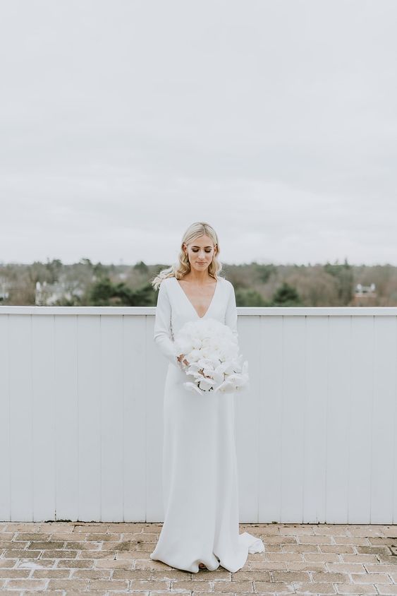 a pretty and simple modern winter bridal look with a plain wedding dress with a depe V-neckline, long sleeves and a train plus a lush white wedding bouquet