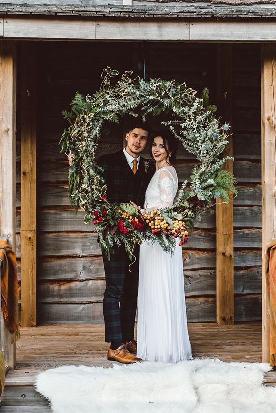 a small yet pretty round wedding arch of greenery, evergreens, red blooms and berries is a gorgeous idea for your Christmas ceremony