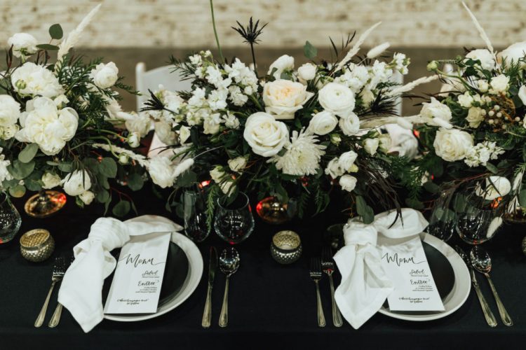 a bold modern winter wedding tablescape with a black tablecloth and plates, with neutral napkins and menus plus neutral blooms