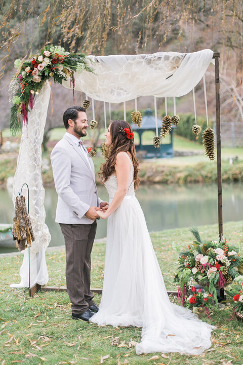 a creative Christmas wedding arch covered with tulle, with evergreen, blush and red flower arrangements and with large pinecones hanging down