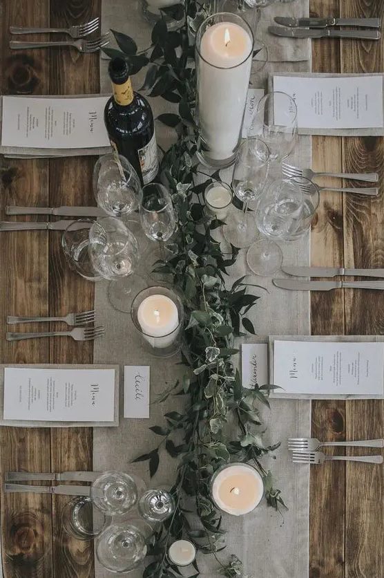 a chic winter wedding tablescape with a grey table runner and linens, candles, a greenery runner and elegant cutlery