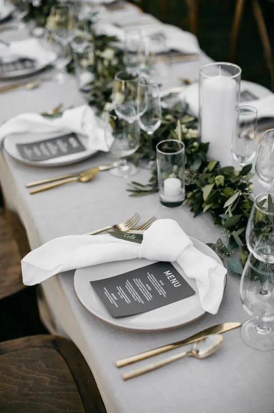 a modern refined wedding tablescape with green and white linens, a greenery runner, candles, gold cutlery is amazing