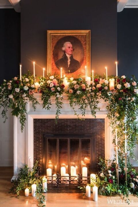 a gorgeous fireplace with pillar candles, cascading greenery, blush, white and burgundy blooms and thin candles is a lovely Christmas wedding backdrop