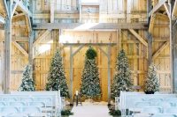31 a white barn wedding ceremony space with several Christmas trees with lights and decor, white chairs and evergreens to line up the aisle
