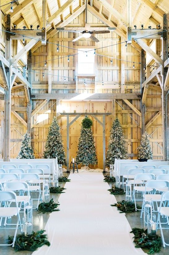 a white barn wedding ceremony space with several Christmas trees with lights and decor, white chairs and evergreens to line up the aisle