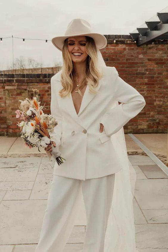 a beautiful and chic 70s inspired bridal look with a white pantsuit with an oversized blazer, layered necklines, a creamy hat with a veil