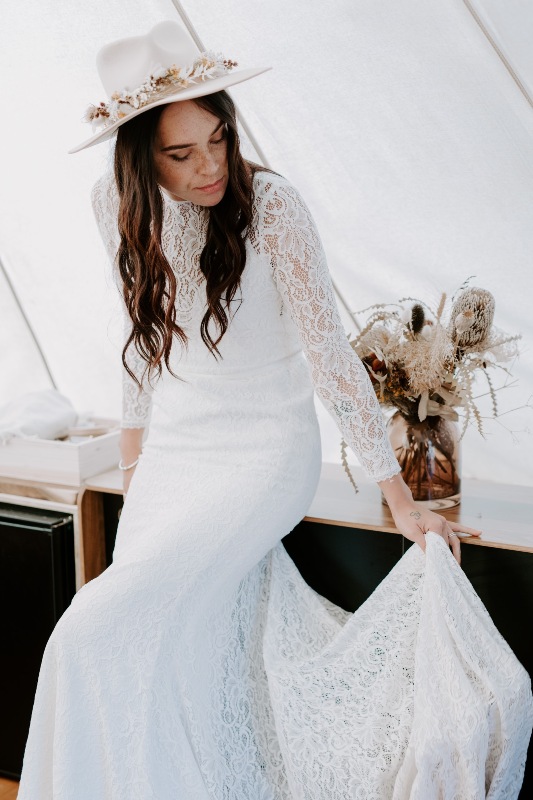 a boho bride wearing a boho lace fitting wedding dress with long sleeves and a high neckline, creamy hat with dried blooms and grasses