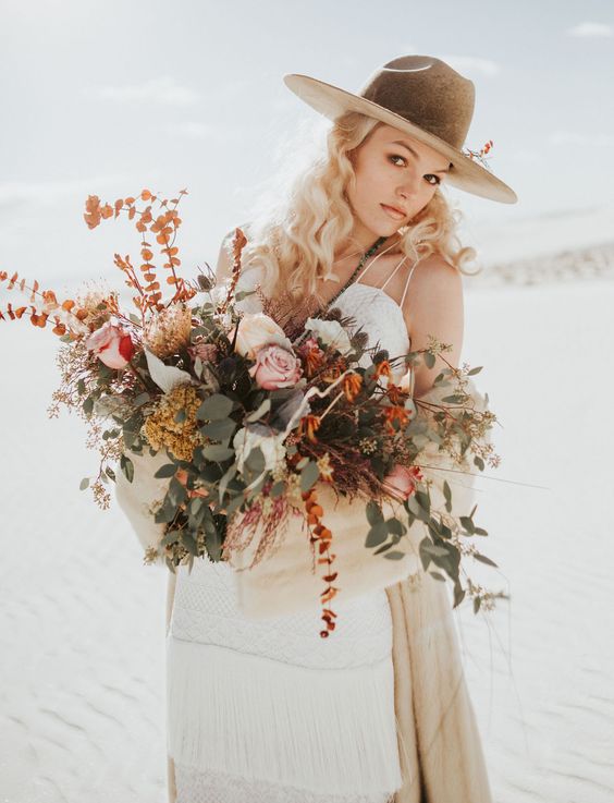 a boho desert bride wearing a fringe tiered wedding dress, a greige hat with some bold foliage and a neutral coverup