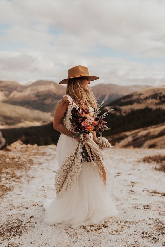a boho western bridal look with an A-line wedding dress with a tulle skirt, a deep neckline and no sleeves, a beige hat and a cool colored bouquet