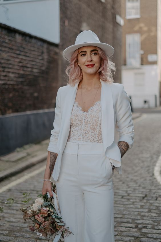 a bride wearing a white pantsuit with a cropped blazer, a white lace bodice and a white fedora hat plus pink hair looks fantastic