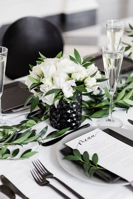a chic modern wedding centerpiece of a black geometric vase, white blooms and greenery and a greenery table runner is elegant