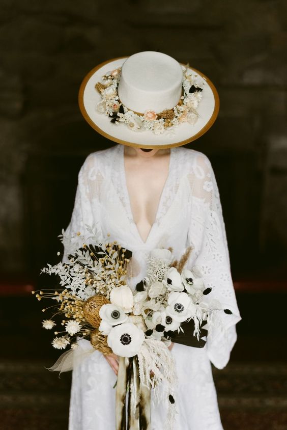 a gorgeous boho bridal look with a white lace wedding dress with a plunging neckline, a white hat with gold rims and neutral blooms