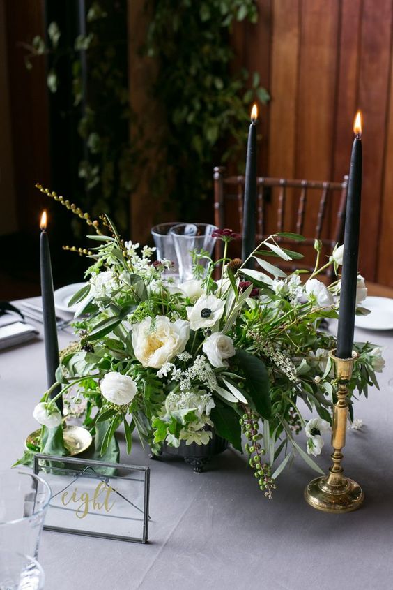 a lovely wedding centerpiece of white blooms and greenery and tall and thin black candles in refined gold candleholders is amazing