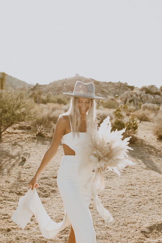 a modern boho bridal look with a plain fitting wedding dress with cutouts, a grey embellished aht and a cool dired weddin bouquet
