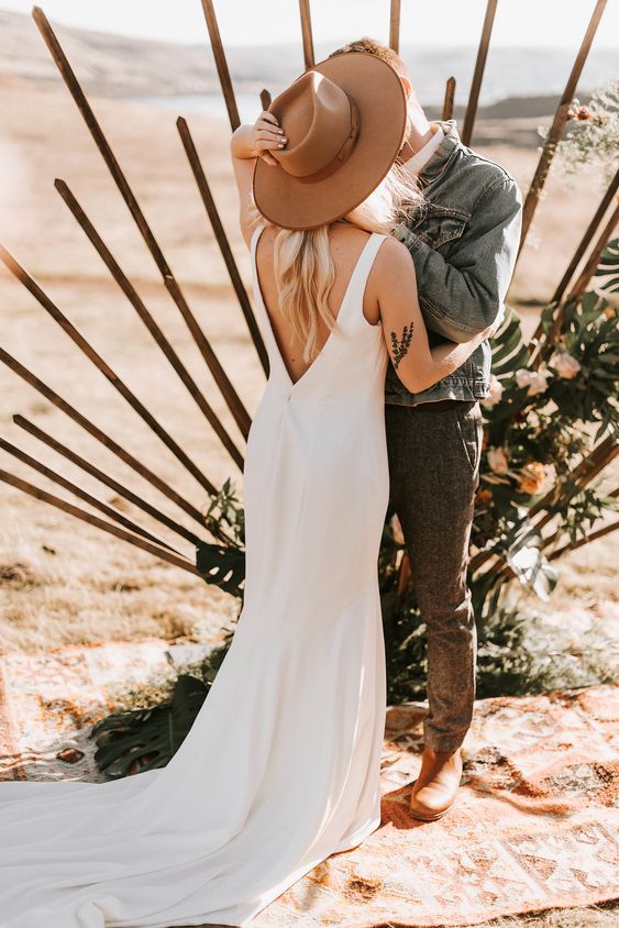 a modern desert bridal look with a plain mermaid wedding dress with a train and a cutout back, a taupe hat for a creative touch