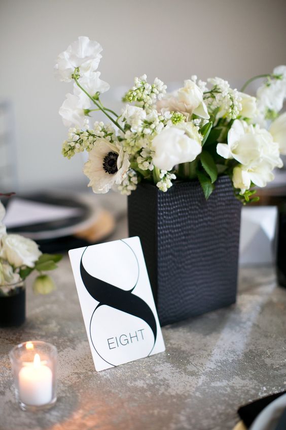 a pretty modern wedding centerpiece of a textural black vase, white blooms and greenery and a table number plus a candle is wow