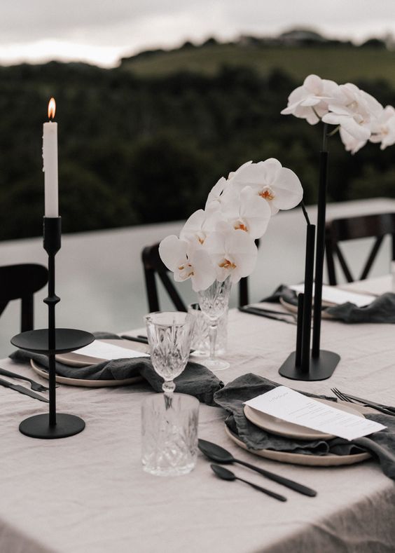 a refined and chic wedding centerpiece of white orchids and black candleholders plus thin and tall white candles is a lovely idea