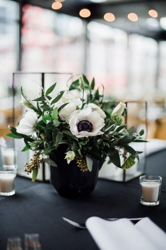 a stylish black and white wedding centerpiece of a black vase, anemones, greenery and some white candles around is pure elegance