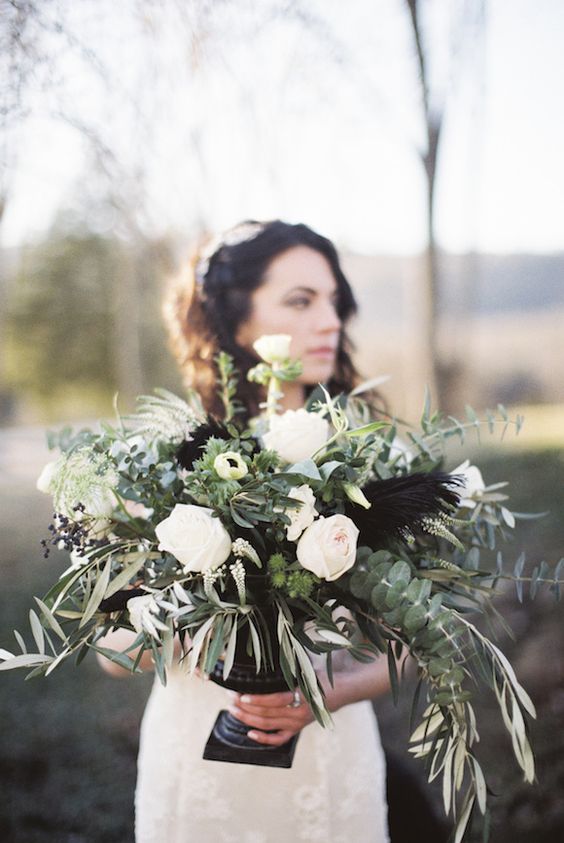 a stylish wedding centerpiece of white blooms, lots of greenery and black feathers plus a black urn is a chic idea for a modern wedding