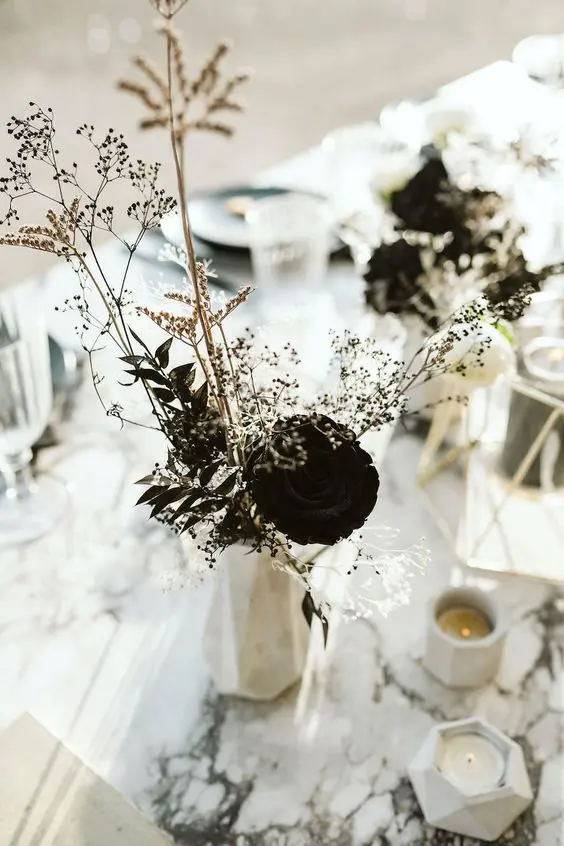an edgy wedding centerpiece of a faceted vase, black blooms and dried leaves and some grass plus faceted candles is a very stylish idea