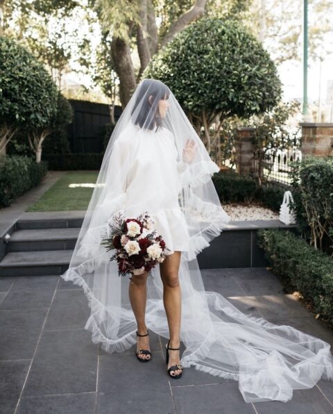 a beautiful A-line oversized wedding dress with a high neckline and puff sleeves, black logo shoes, a long fringe veil inspired by the 70s