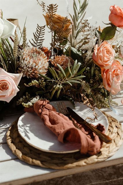 a beautiful copper bronze wedding tablescape with lush florals and greenery, a woven placemat and a rust-colored napkin is chic