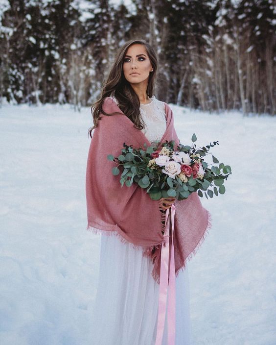 a beautiful winter bridal look with a neutral lace embellished wedding dress, a pink cover up, a neutral and pink bouquet with pink ribbons