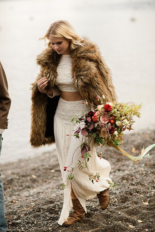 a boho bridal look with a two-piece wedding dress with lace, brown boots and a brown fur coat plus a lush and bold wedding bouquet