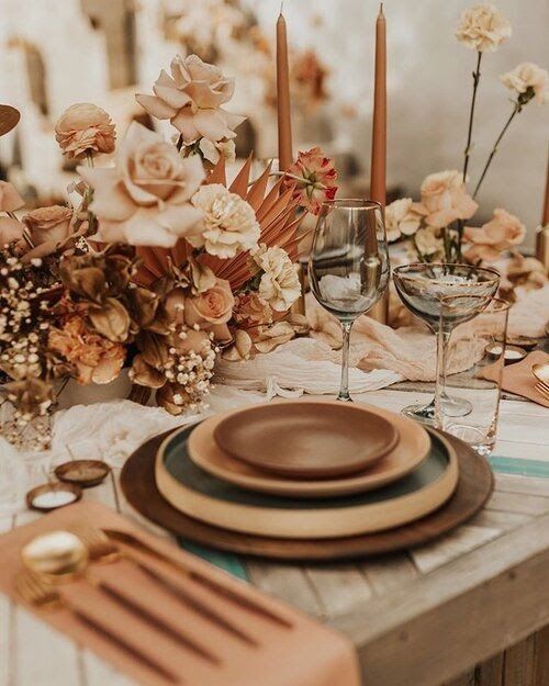 a refined and beautiful boho earthy wedding tablescape with a wooden placemat and brown and amber plates, rust and peachy blooms, copper candles and napkins