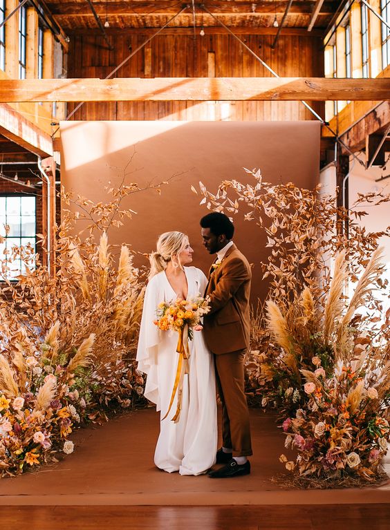 an earthy-toned floral wedding altar with blooms, grasses and dried leaves, a rust-colored backdrop is amazing