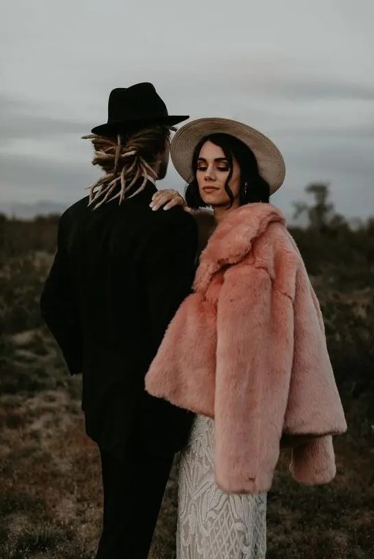 a gorgeous pink faux fur coat over a romantic boho lace wedding dress plus a hat for a boho bridal look in fall or winter