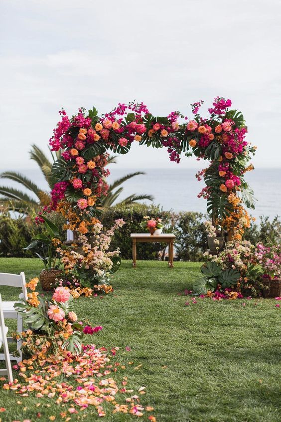 a super colorful wedding arch with tropical leaves, pink, orange, yellow and neutral blooms all over for a tropical wedding