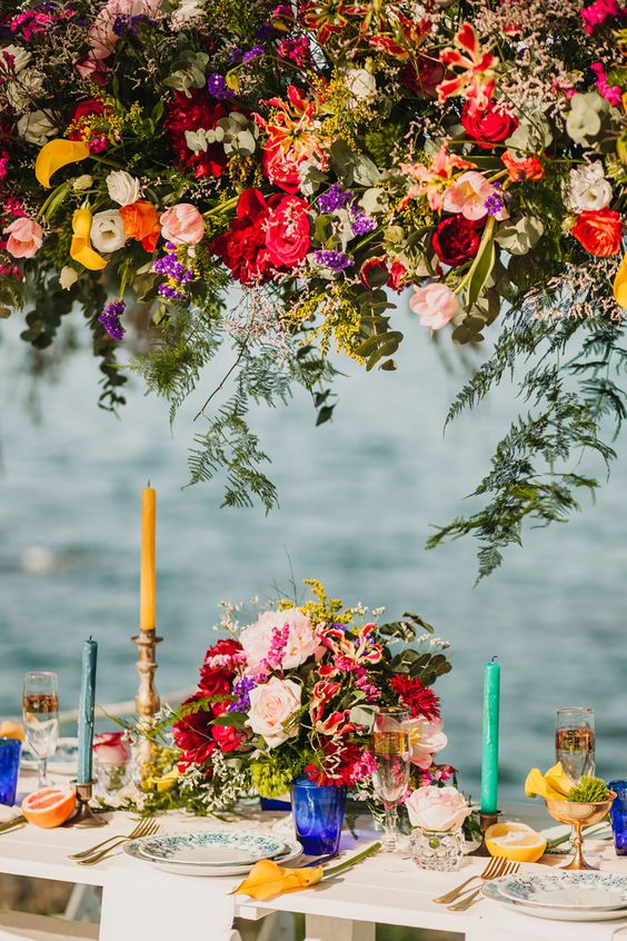 a super bright wedding reception with bold florals and greenery over the table and on the table, colorful candles and glasses