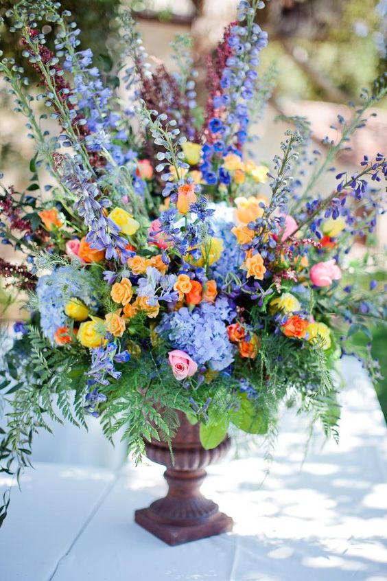 a colorful wedding floral arrangement of a vintage urn with orange, yellow, pink, periwinkle blooms and lots of greenery