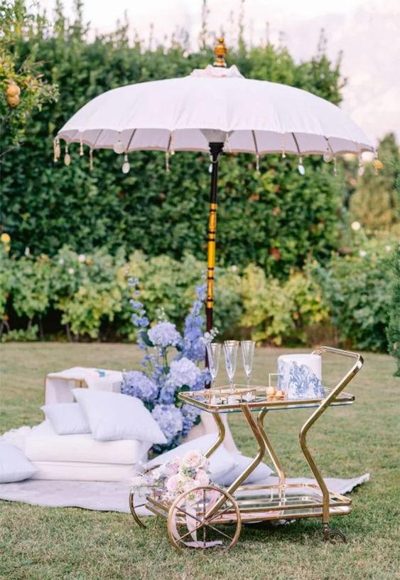 a cozy wedding nook with a blush umbrella, lilac pillows and blankets, blue blooms, a cart with glasses and a periwinkle decorated wedding cake