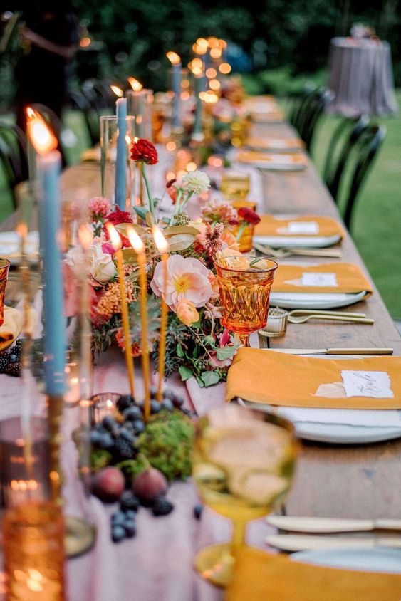 a super colorful wedding table setting with yellow napkins and a pink runner, blue candles, bold and lush florals