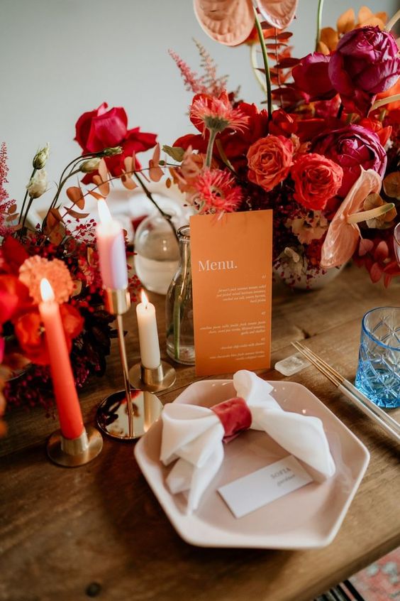 a bright and chic wedding tablescape with red, orange and pink blooms, an orange menu, colorful candles and a pink hex plate