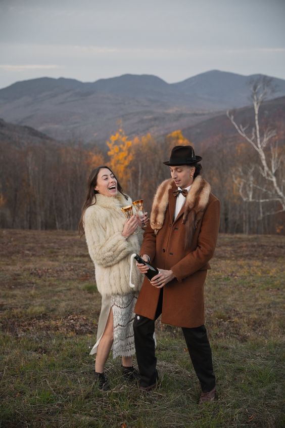 a creative boho groom's look with a rust-colored coat with a fur collar, black trousers, brown shoes and a brown bow tie