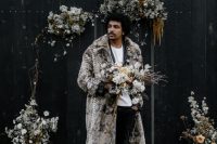 a modern groom’s look with a black velvet pantsuit, a white t-shirt, black shoes and a greige faux fur coat with a print is amazing for a winter affair